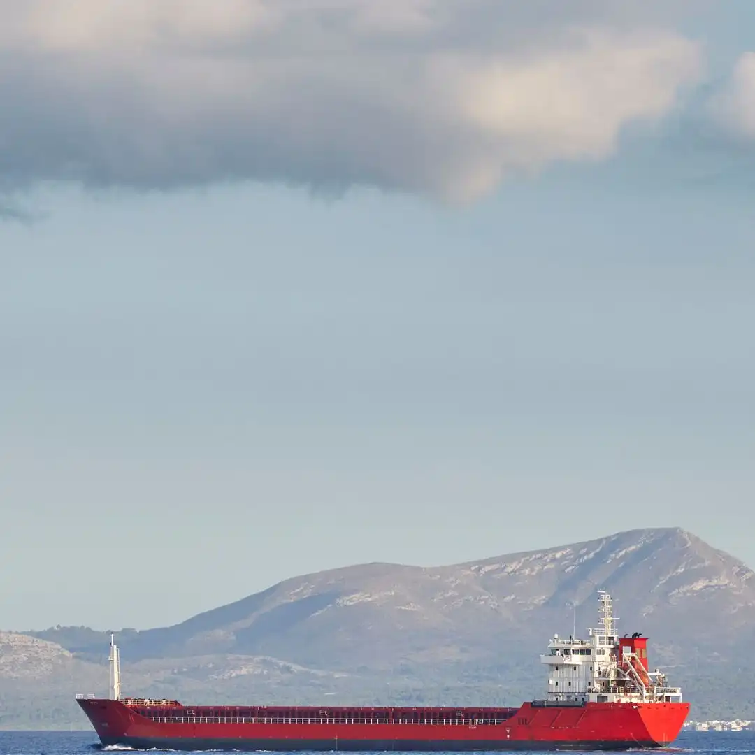 A red bulk carrier underway on calm water with a mountainous coastline behind