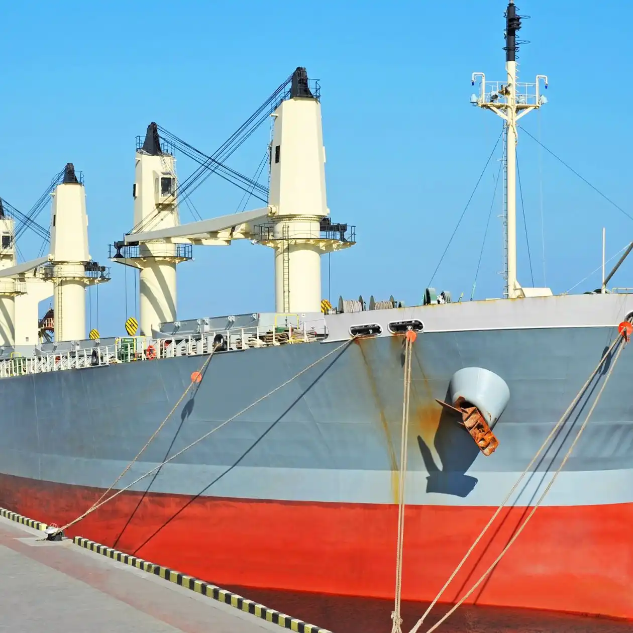 A geared bulk carrier with deck cranes moored at a concrete pier