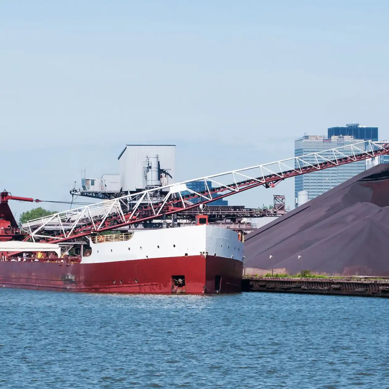 A red bulk carrier handling iron ore via a shore conveyor at a Brazilian terminal